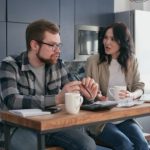 Adult couple sitting at a table. He is wearing a check shirt with various shades of grey and has a beard and glasses. She is talking to him. She has medium long dark hair, a white top and a beige shirt over the top.