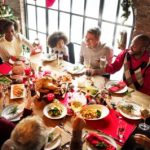 Large family group with black and white adults and children seated around a table with a Christmas dinner on it. The room has Christmas decorations.