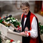 Bishop Mariann Edgar Budd. Mature white woman with short grey hair at a pulpit wearing a white robe with red vestment and a black sash