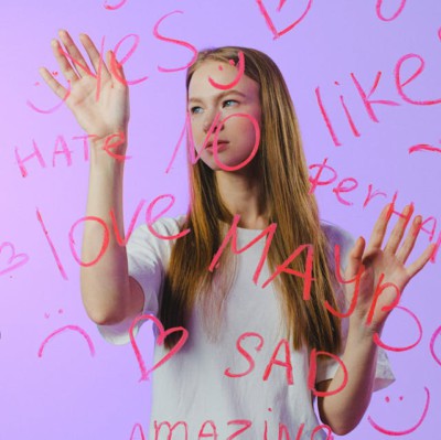 Teenage girl with long blonde hair putting her hands onto a glass screen with various words written in red. Emotional words such as love, like, amazing, happy and sad. Smiley faces and hearts
