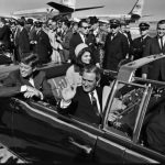 Black and white image of John F Kennedy in the back of an open top car at Dallas airport with Jackie Kennedy seated next to him