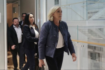 Marie Le Pen walking out of a door followed by three others in suits. She is a mature woman with shoulder length blonde hair, wearing a blue jacket with a white top and black trousers.