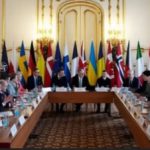 Various country leaders seated around a table with the nation flags in the background