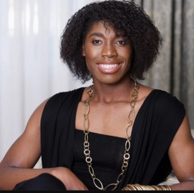 A black adult woman with slightly long hair smiling. She is seated wearing a black dress with a necklace.