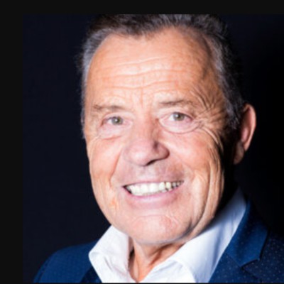 Appearance of a mature man with thin dark hair wearing a blue jacket and white shirt.