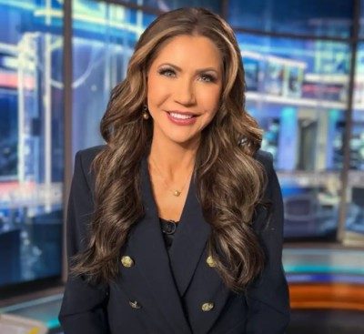 Kristi Noem. Adult white woman with long dark hair. Wearing a dark jacket with large brass buttons.
