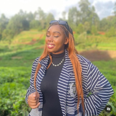 A black woman with long straightened hair coloured ginger. Wearing a blue and white striped jacket, black top and necklace. Standing in a blurred tree and fields location.