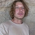 Mykhailo Viktorovych Polyakov. A white young man with longish hair and a bit of face fluff. Wearing a white top posed against a rocky background. Possibly limestone.