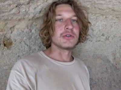 Mykhailo Viktorovych Polyakov. A white young man with longish hair and a bit of face fluff. Wearing a white top posed against a rocky background. Possibly limestone.