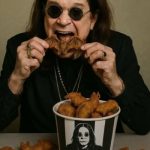 Ozzy Osbourne eating a breadcrumb coated item in the shape of a bat. There are various other breadcrumb coated bats on the table in front of him. There is also a large bucket of bats. The bucket has black vertical stripes with a stylised image of Ozzy Osbourne and the writing, SFB. Sabbath Fried Bats