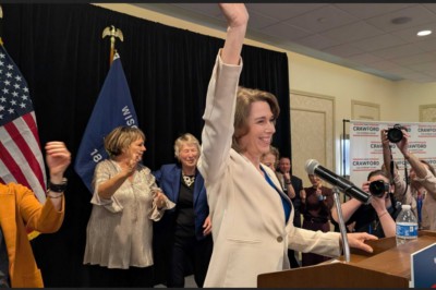 Susan Crawford celebrating her victory. A mature white lady with shoulder length light brown hair. Wearing a cream coloured jacket. Several people around her applauding.