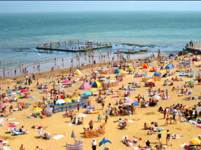 UK beach scene in summer. A yellow sandy beach with lots of people with seaty, umbrellas and wind breaks. People are playing in the blue sea in the distance. Broadstairs in Kent.