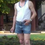 Old man in shorts. White man with glasses and thinning hair wearing blue denim shorts and a white vest standing in front of a lawn with a house in the background.