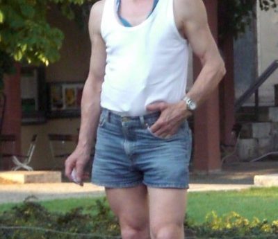 Old man in shorts. White man with glasses and thinning hair wearing blue denim shorts and a white vest standing in front of a lawn with a house in the background.