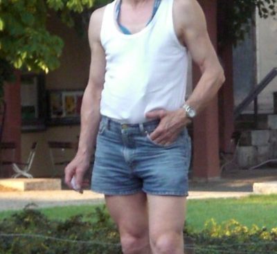 Old man in shorts. White man with glasses and thinning hair wearing blue denim shorts and a white vest standing in front of a lawn with a house in the background.