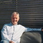 A mature white man with short grey hair in a white shirt and jeans sitting on a grey sofa with grey Venetian blinds behind him