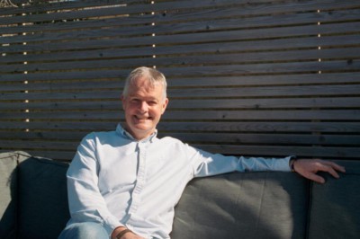 A mature white man with short grey hair in a white shirt and jeans sitting on a grey sofa with grey Venetian blinds behind him