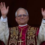 Pope Leo XIV. White man with short receding grey hair wearing glasses. His hands are raised up. He is wearing an embroidered white robe with a red over garment and a heavily embroidered sash. A large gold cross hangs from his neck.