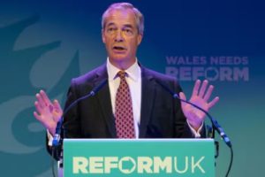 Nigel Farage, knob head in a black suit, white shirt and a red tie standing at a podium behind a sign 'ReformUK'