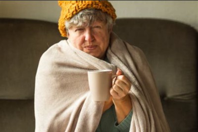 Old white lady wrapped in a blanket with a ginger woolly hat on holding a mug of drink. She is sitting on a sofa.