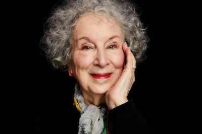 A mature woman with grey curly hair. She is wearing a black top with a light grey scarf. The background is black.