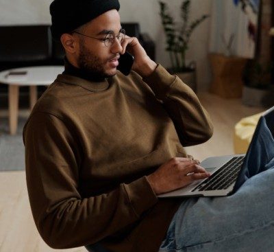 man in brown long-sleeve shirt and jeans sitting on a chair looking at a laptop with a mobile phone to his ear