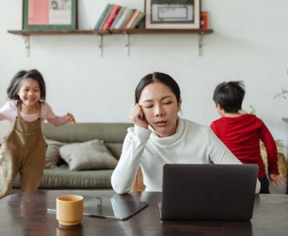 Mother looking at laptop while two young children are running around behind her