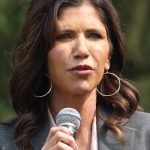 White woman with long wavy brown hair. Wearing a grey jacket and white top. She is holding a microphone.