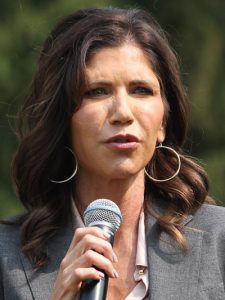 White woman with long wavy brown hair. Wearing a grey jacket and white top. She is holding a microphone.