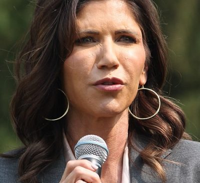 White woman with long wavy brown hair. Wearing a grey jacket and white top. She is holding a microphone.