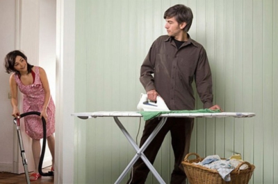 A woman in a pink dress is vacuum cleaning the floor while a man is ironing