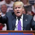 Donald Trump speaking at a podium with his right hand raised and pointing. He.is wearing a dark navy blue suit, white shirt and purple tie.