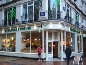A Starbucks coffee shop. A white corner building with large windows on either side. One person standing outside while another is walking away.