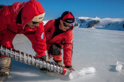 Two climate scientists in red outdoor insulated jackets in an icy landscape with a large augur drill collecting ice core samples.
