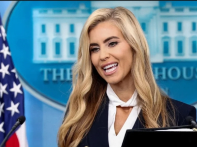 Bottle blonde bimbo Kate Zachariah standing in front of the White House backdrop.